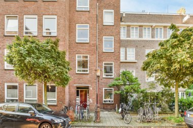 Amsterdam, Hollanda - 10 Nisan 2021: tuğladan bir apartmanın önüne park edilmiş iki araba, yanında bisikletler ve birkaç ağaç var.