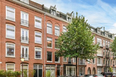 Amsterdam, Hollanda - 10 Nisan 2021: kaldırımlara park etmiş arabaların ve kırmızı tuğlalı apartmanların önündeki ağaçların olduğu bir şehir caddesi.