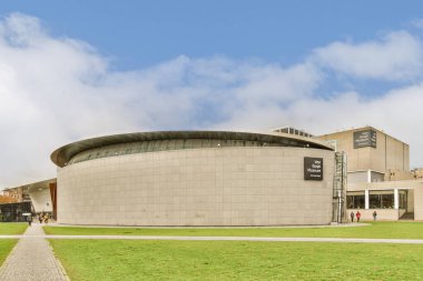 Çimenli bir alanın ortasında bir bina. Etrafında insanlar yürüyor ve her iki tarafında binalar var.