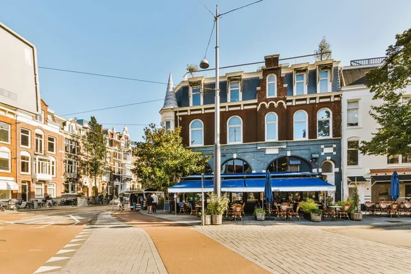 stock image Amsterdam, Netherlands - 10 April, 2021: a city street with buildings in the background and people sitting at tables under umbrellas on either side of the street