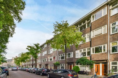 Amsterdam, Hollanda - 10 Nisan 2021: bazı arabalar yeşil ağaçlı tuğla apartman binalarının önünde caddenin kenarına park edilmiş.