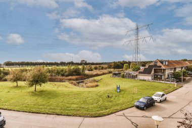 Arazinin ortasında bir ev. Garaj yoluna park edilmiş arabalar ve elektrik hatları avludan geçiyor.