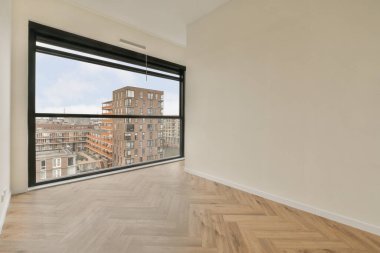 Boş bir oda, ahşap döşemeli ve geniş pencereli. Şehir manzarasının içinden çekilmiş bir fotoğraf.