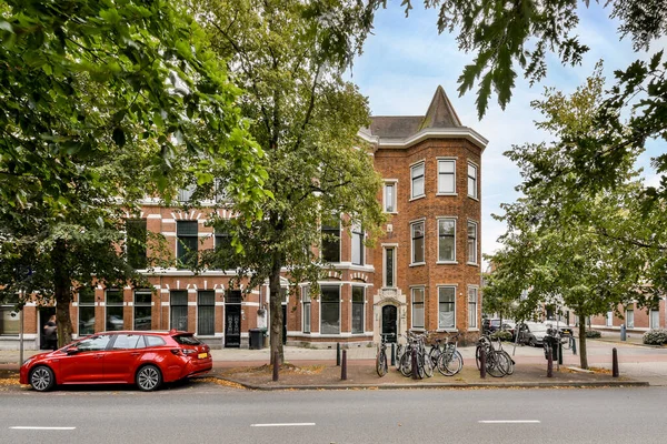 stock image Amsterdam, Netherlands - 10 April, 2021: a red car parked in front of a large brick building with a clock tower on the top and two bicycles attached to its