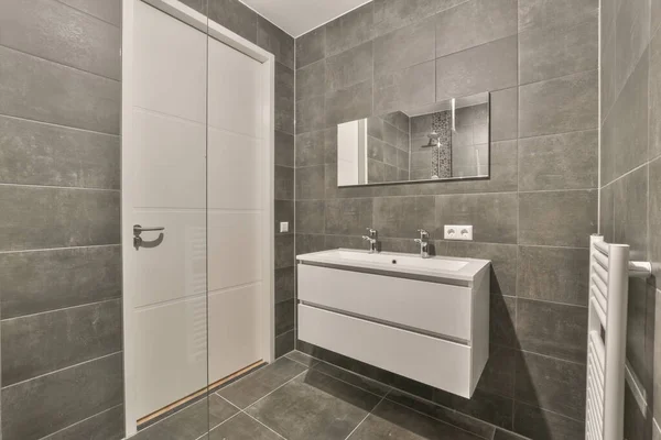 壁にグレーのタイルと白い器具を備えたモダンなバスルームシンクとシャワーエリアのドアの上に鏡 — ストック写真