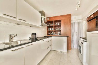Görüntünün ortasında beyaz dolapları ve siyah tezgahları olan modern bir mutfak bir fırın, bulaşık makinesi ve lavabo.