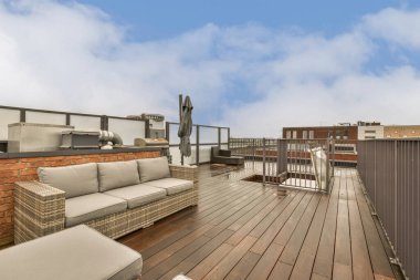 Mavi gökyüzünde ahşap döşemeli ve beyaz bulutlu bir açık hava yaşam alanı sağ tarafta görülebilir.