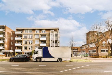 Mavi gökyüzü ve beyaz bulutlu bir apartmanın önünde yolun kenarına park etmiş hareket eden bir kamyon.