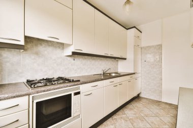 Bu fotoğraftaki tezgahların üzerinde beyaz dolaplar ve mermer tezgahlı bir mutfak ön taraftan çekiliyor.