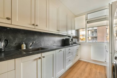 Bu fotoğrafta tezgahların üzerinde beyaz dolaplar ve siyah tezgahlar olan bir mutfak içeriden çekilmiş.