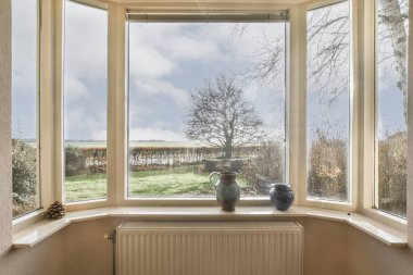 Pencere kenarında vazo olan bir pencere ve ağaçların arasından bir tarla manzarası.