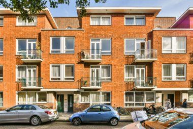 Amsterdam, Hollanda - 10 Nisan 2021: Birçok pencere ve balya bulunan bir apartmanın önündeki yolun kenarına park edilmiş bazı arabalar