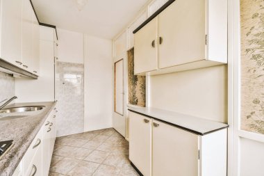 Bu fotoğrafta yerde beyaz dolapları ve mermer tezgahları olan bir mutfak alanı içeriden çekilmiş.