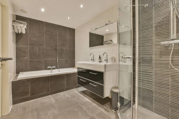 stock image a modern bathroom with grey tiles and white fixtures in the shower stall, toilet and bathtub are visible on the wall