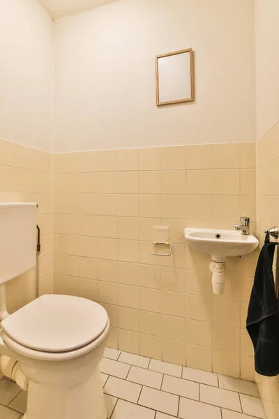 一个白色的厕所和一个小浴室的水池 墙壁上有米黄色的瓷砖 还有一条黑色的毛巾 — 图库照片