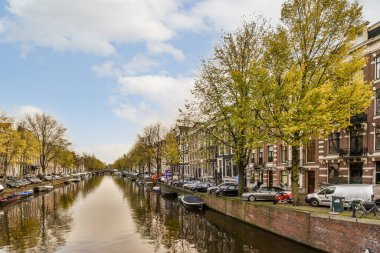 Amsterdam, Hollanda - 10 Nisan 2021: Güneşli bir günde önü kayık ve ağaçlarla dolu bir kanal, suların ötesinden görüldüğü gibi.