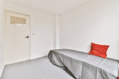 Beyaz duvarlı ve gri halılı bir yatak odası. Yatağın üzerinde kırmızı bir yastık var.