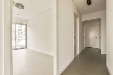 Beyaz duvarları ve gri zemini olan boş bir oda. Başka bir odaya açılan bir kapı var.