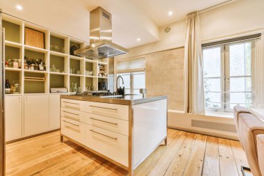 Ahşap döşemeli ve arkasındaki duvarda beyaz dolaplı bir mutfak sokağa bakan büyük bir penceredir.