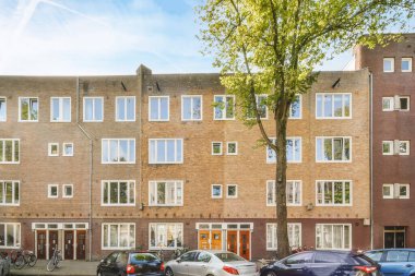 Amsterdam, Hollanda - 10 Nisan 2021: Bazı arabalar binanın üst katında ve alt yarısında birçok penceresi olan büyük tuğla bir binanın önüne park ettiler.