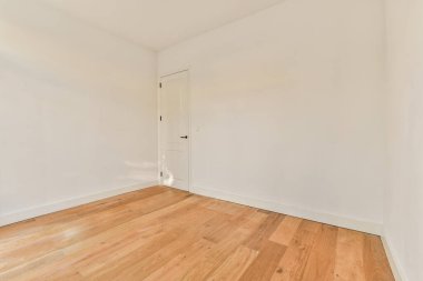 Beyaz duvarları ve sağ tarafında ahşap döşemeleri olan boş bir oda, sol köşede bir kapı var.