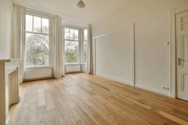 Boş bir oturma odası, ahşap döşeme ve pencereden ağaçlara bakan geniş pencereli.