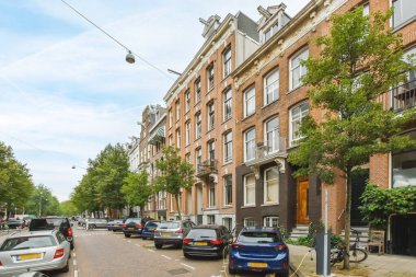 Amsterdam, Hollanda - 10 Nisan 2021: Kenarında arabalar olan bir şehir caddesi ve bazı apartmanların önünde kaldırımdan yürüyen insanlar