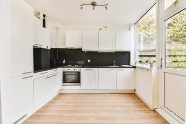 Açık bir pencerenin önünde, üzerinde beyaz dolaplar ve siyah tezgahlar olan bir mutfak ağaçlara bakıyor.