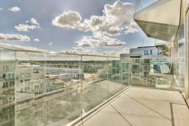 Gökyüzünde binalar ve bulutlar olan bir dış alan. Şehir manzaralı bir balkonda cam korkuluklardan görüldüğü gibi.