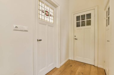 Kapının her iki tarafında beyaz kapılı ve ahşap döşemeli boş bir oda, duvarda bir pencere var.