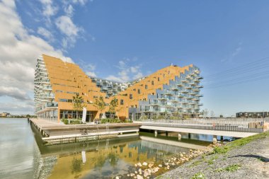 Turuncu bir binanın önündeki su kütlesinin yanındaki bazı binalar ve beyaz bulutlu mavi gökyüzü.