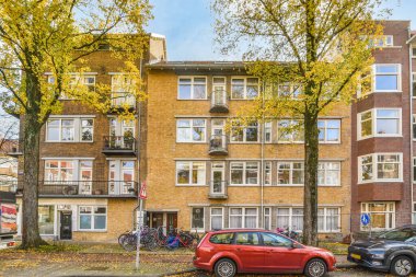 Amsterdam, Hollanda - 10 Nisan 2021: Yanı başında sarı yapraklar olan bir apartmanın önüne park edilmiş iki araba kırmızı bir araba.
