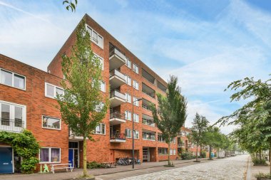 Amsterdam, Hollanda - 10 Nisan 2021: Her iki tarafında ağaçlar ve yol boyunca park edilmiş arabalar olan tuğla bir binanın önündeki boş bir cadde