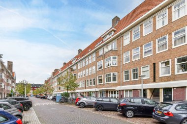Amsterdam, Hollanda - 10 Nisan 2021: Beyaz pencereli ve kırmızı çatılı tuğla bir binanın önündeki caddenin kenarına park edilmiş bazı arabalar