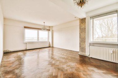 Boş bir oturma odası, ahşap döşeme ve pencereden ağaçlara bakan geniş pencereli.