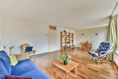 Odada mavi kanepesi ve ahşap sehpası olan bir oturma odası beyaz duvarlar, ahşap döşemeler ve büyük pencereler var.