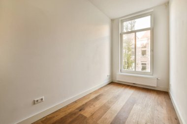 Ahşap döşemeleri ve beyaz duvarları olan boş bir oda, sağdaki duvarda bir pencere var.