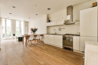 Ahşap zemin, beyaz dolaplar, paslanmaz eşyalar ve verandaya açılan açık bir kapı olan bir evde mutfak ve yemek odası.