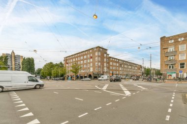 Açık mavi bir gökyüzünde, bir apartmanın önünde, yoldan geçen arabaları ve otobüsleri olan bir cadde.