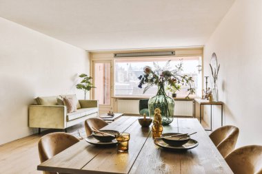 Bir yemek masası ve büyük pencereli bir odada aşağıdaki sokağa bakan bir vazo dolusu çiçek var.