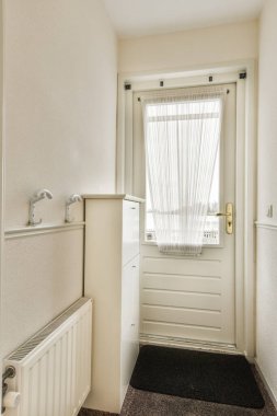 Duvarlarında beyaz süslemeler olan küçük bir odada bir kapı ve pencere, halı kaplı zemin ve ışık saçan