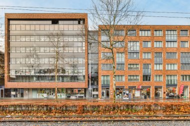 Amsterdam, Hollanda - 10 Nisan 2021: Birçok pencere ve ağacın bulunduğu tuğla bir binanın önündeki tren yolunun kenarındaki bir ofis binası