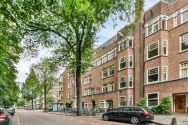 Amsterdam, Hollanda - 10 Nisan 2021: yan tarafında arabaların park ettiği bir sokak ve apartman binalarının önündeki yol boyunca uzanan ağaçlar, kırmızı tuğlalı.