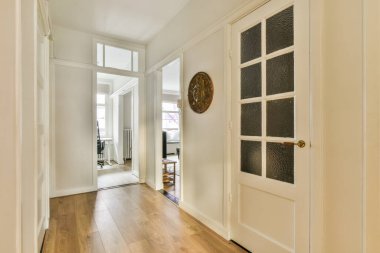 Duvara monte edilmiş antika bir saat tarafından çerçevelenmiş, ahşap döşemeli ve beyaz süslemeli bir koridor.