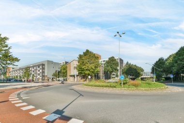 Caddenin ortasında bir kavşak. Yolun karşısından her iki tarafında da binalar ve ağaçlar var.