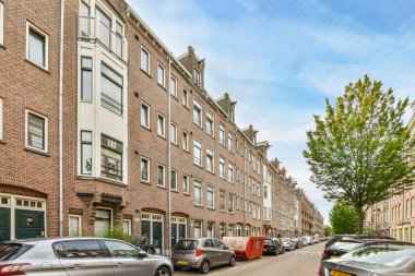 Amsterdam, Hollanda - 10 Nisan 2021: Bir kentsel alanda caddenin kenarına park edilmiş bazı arabalar, her iki tarafında da binalar ve ağaçlar var.