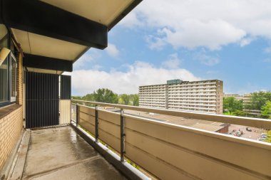 Arka planda bazı binalar ve üzerinde mavi gökyüzü olan bir balkon, bir apartman binasının balkonundan görüldüğü gibi.