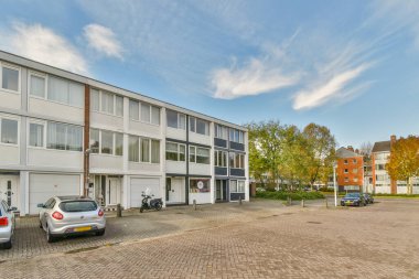 Bir apartman kompleksi. Garaj yolunda arabaları ve caddeden görüldüğü üzere binanın her iki tarafında da ağaçları olan bir bina.