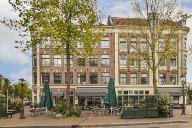 Amsterdam, Hollanda - 10 Nisan 2021: Her iki tarafında birçok masa, sandalye ve şemsiye bulunan bir bölgenin ortasındaki bazı bina ve ağaçlar