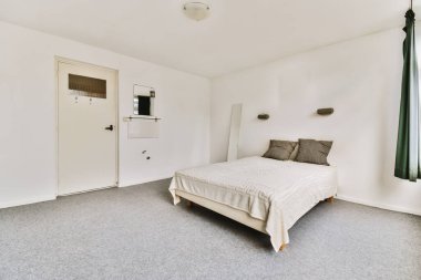 Beyaz duvarları ve gri halıları olan bir yatak odası. Odada küçük bir yatak var. Klimalı ısıtıcı açık.